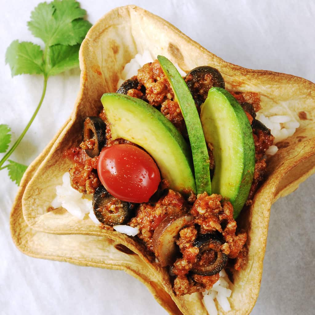 Baked tortilla bowls holding rice, a meat mixture, tomatoes and avocado slices