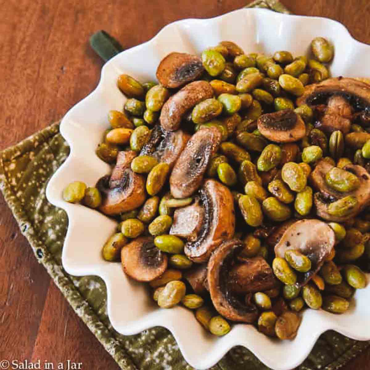 edamame and mushrooms in a serving dish on top of a hot pad.