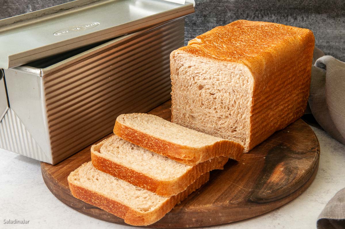 partially sliced honey wheat loaf sitting next to a Pullman pan
