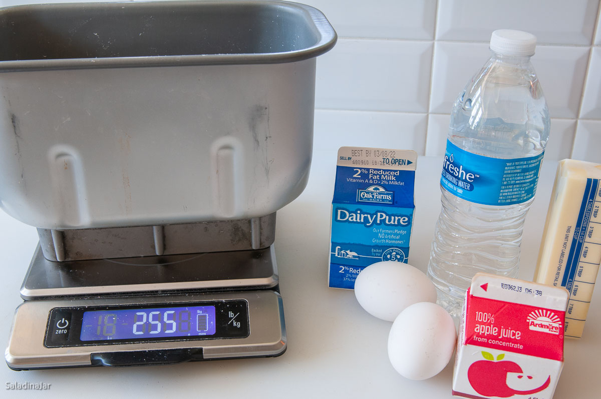 bread machine pan on scales next to liquids including eggs, butter, and fruit juice.