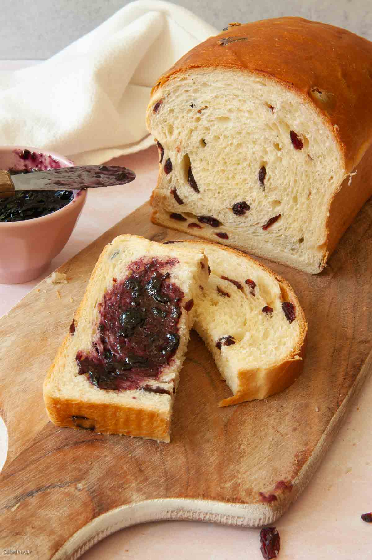 Sliced dried fruit bread on a cutting board-- one piece slathered with blackberry jelly.