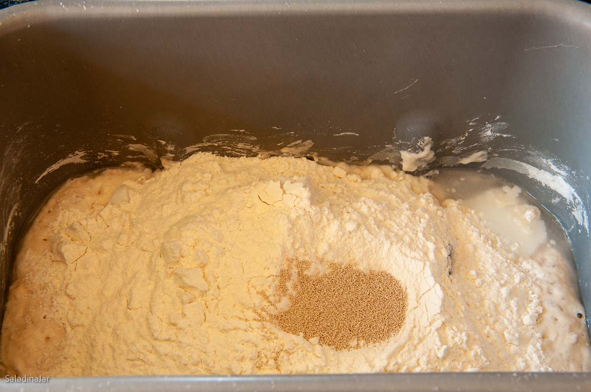 Combining the preferment and remainder of the dough ingredients in a bread machine pan.